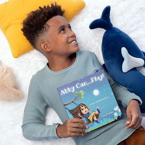 “A young child lies on a soft white surface, smiling while holding a children’s book titled ‘Abby Can… Play! A story about inclusion.’ A yellow pillow and two plush toys—a whale and a beige animal—surround the child.”