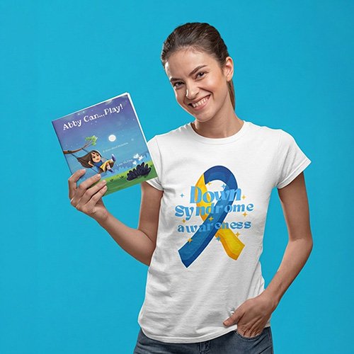 “A person stands against a solid blue background, smiling and holding the children’s book ‘Abby Can… Play!’ They wear a white T‑shirt featuring a blue and yellow ribbon and the words ‘Down Syndrome awareness.’”