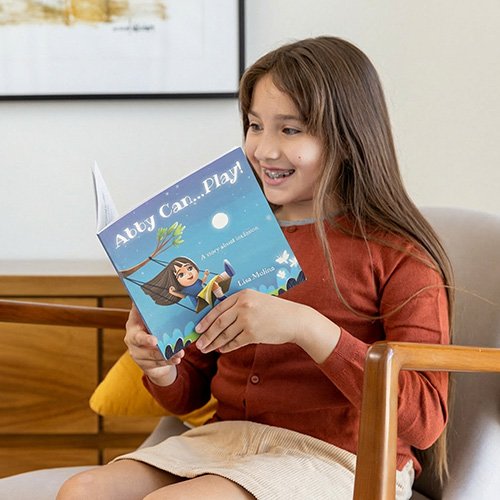 “A young girl sits in a modern armchair, smiling as she reads the children’s book ‘Abby Can… Play!’ A wooden cabinet and a yellow pillow are visible in the cozy living room behind her.”