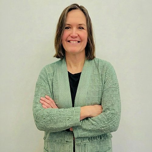 LISA PROFILE WEB A photograph of a smiling woman with shoulder-length brown hair, wearing a light green textured cardigan over a black top. She stands with her arms crossed against a plain white background.